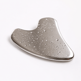 A polished silver metal gua sha massage tool with a wavy, fluid shape and small water droplets on its surface, photographed on a clean white background.