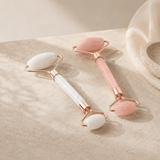 Two rose gold double-ended facial massage rollers with smooth stone heads, one white and one pink, resting on a textured beige surface.