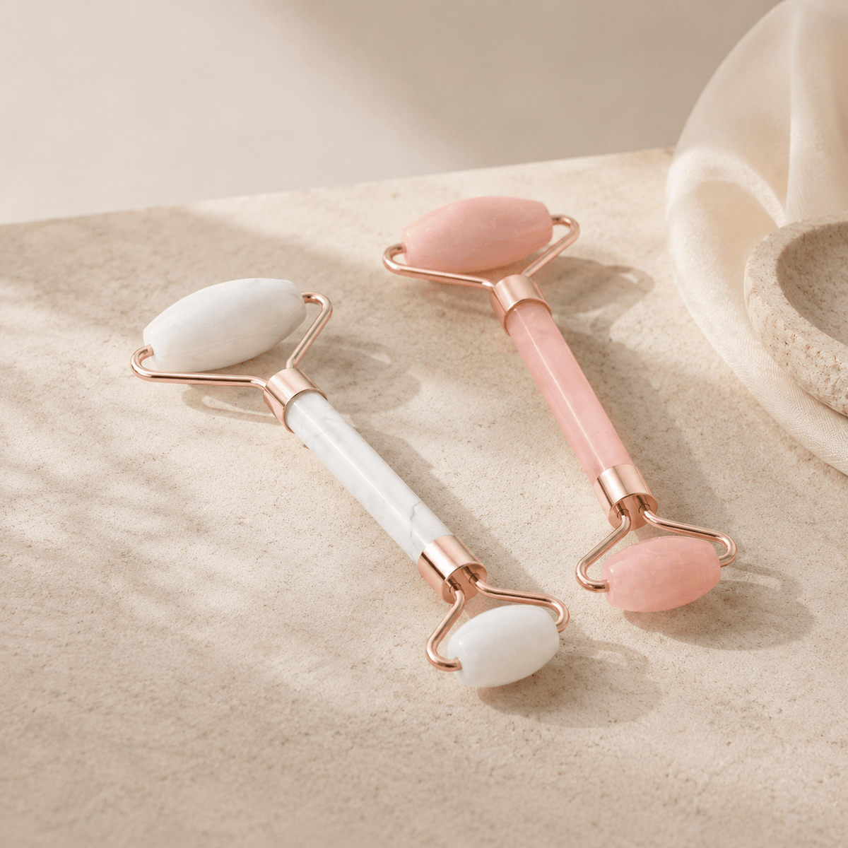 Two rose gold double-ended facial massage rollers with smooth stone heads, one white and one pink, resting on a textured beige surface.