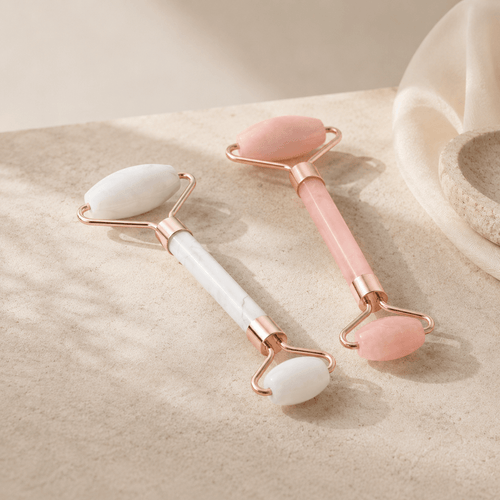 Two rose gold double-ended facial massage rollers with smooth stone heads, one white and one pink, resting on a textured beige surface.